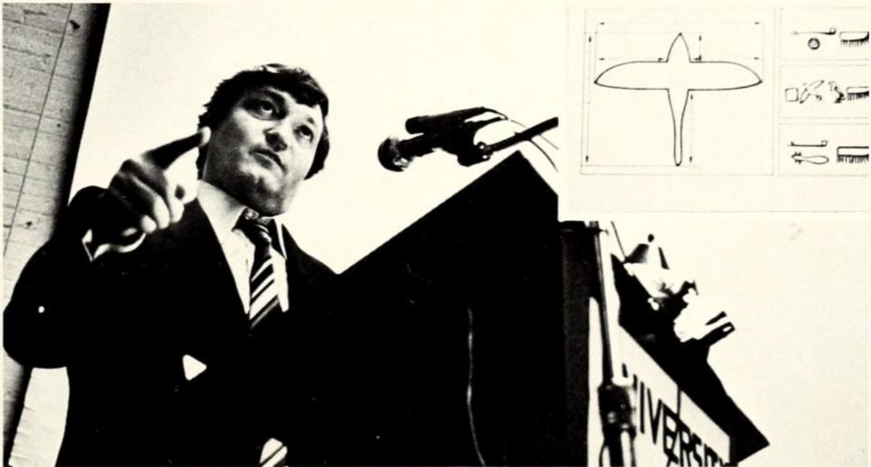 Erich von Däniken at a lectern, pointing, with a drawing of a winged figure projected behind him.